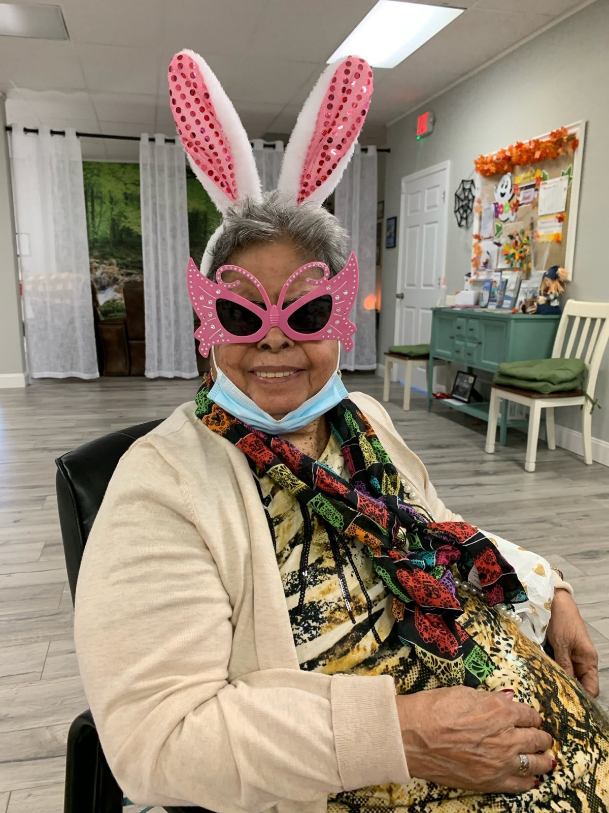An elderly woman wearing bunny ears and pink glasses, sitting indoors with a playful expression.