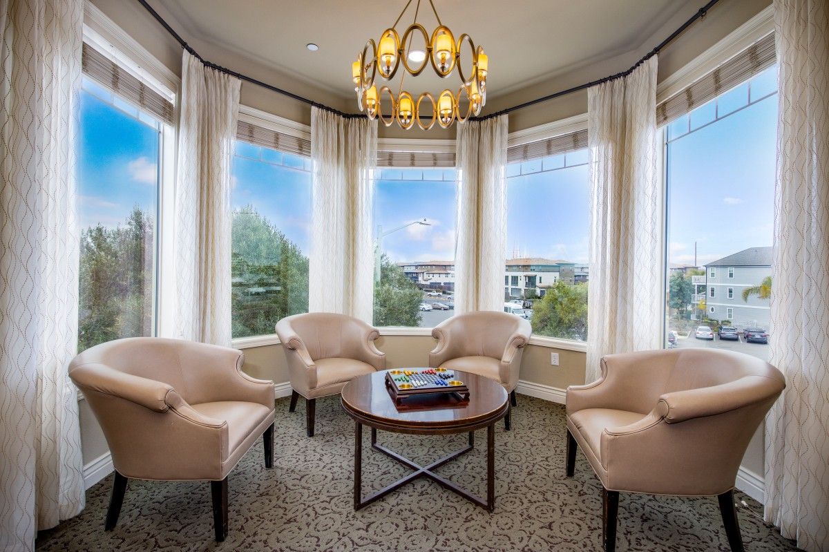 A cozy living room with armchairs arranged around a coffee table, illuminated by natural light through large windows and a central chandelier.
