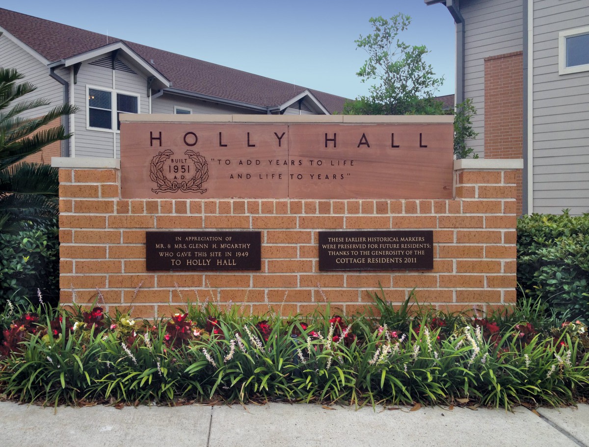 A brick sign for Holly Hall with plaques and flowers in front, located near residential buildings.