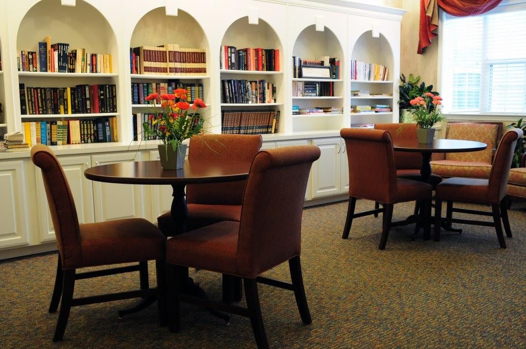 A cozy library space featuring shelves of books, comfortable chairs, and round tables adorned with flowers.