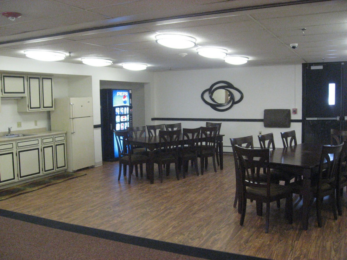 A dining area with multiple tables and chairs, adjoining a small kitchenette, featuring wooden flooring and wall art.