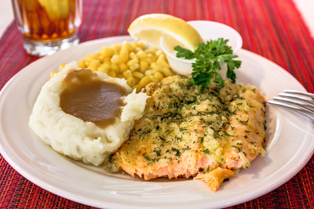 A plate of herb-crusted salmon served with mashed potatoes and gravy, corn, and a lemon wedge.