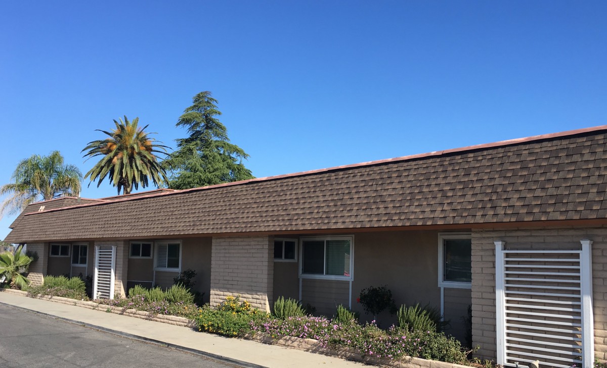 A single-story residential building with a shingled roof, surrounded by trees and landscaping under a clear blue sky.