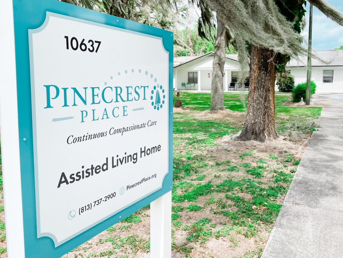 A sign for Pinecrest Place Assisted Living Home is displayed outside, surrounded by trees and grass.
