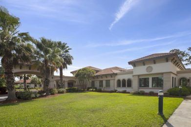 A spacious mansion with a well-maintained lawn and palm trees under a clear blue sky.