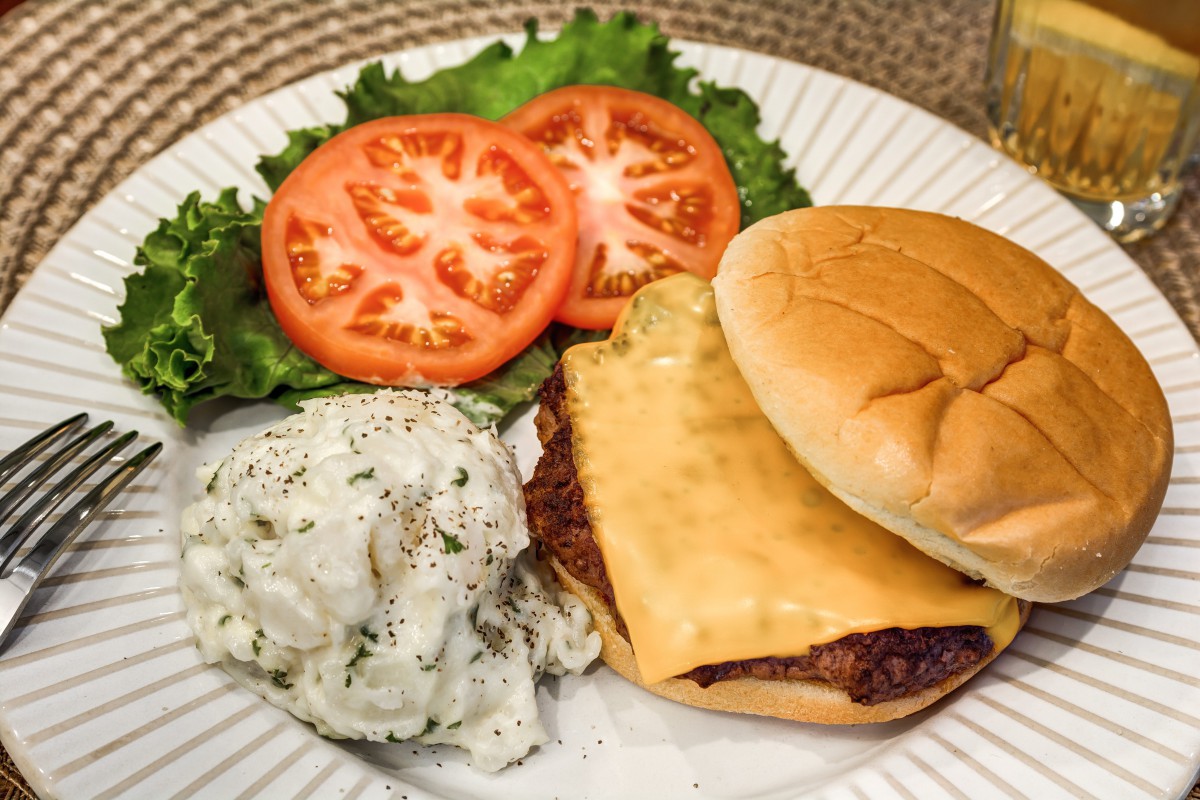 A cheeseburger on a plate with fresh lettuce, sliced tomatoes, and a side of potato salad.