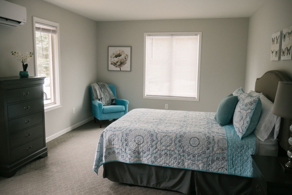 A cozy bedroom featuring a neatly made bed, a turquoise armchair by the window, and soft natural lighting.