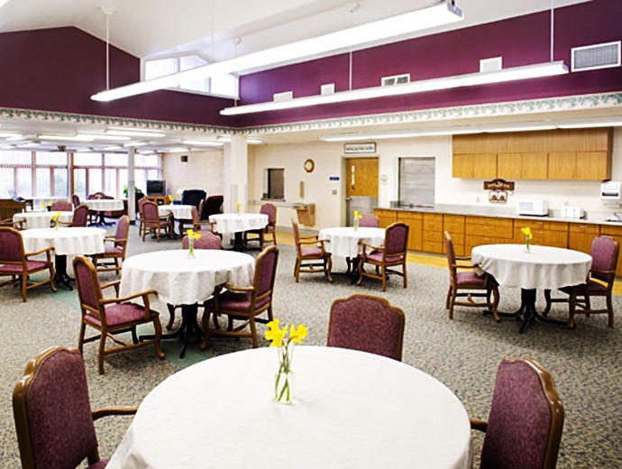 A spacious dining area with round tables covered in white tablecloths and adorned with yellow flowers, featuring a kitchen area in the background.