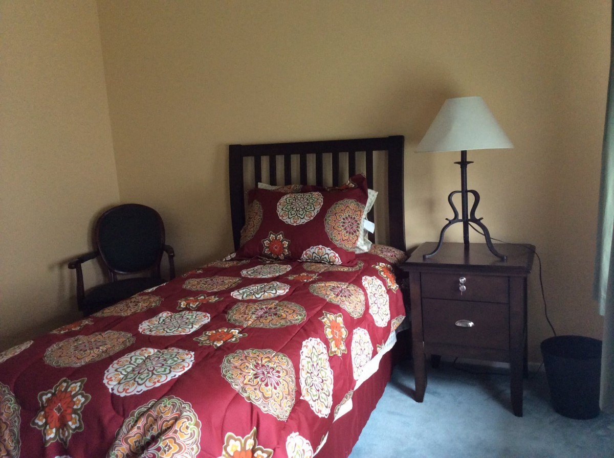 A cozy bedroom featuring a single bed with a colorful quilt, a nightstand with a lamp, and an upholstered chair.