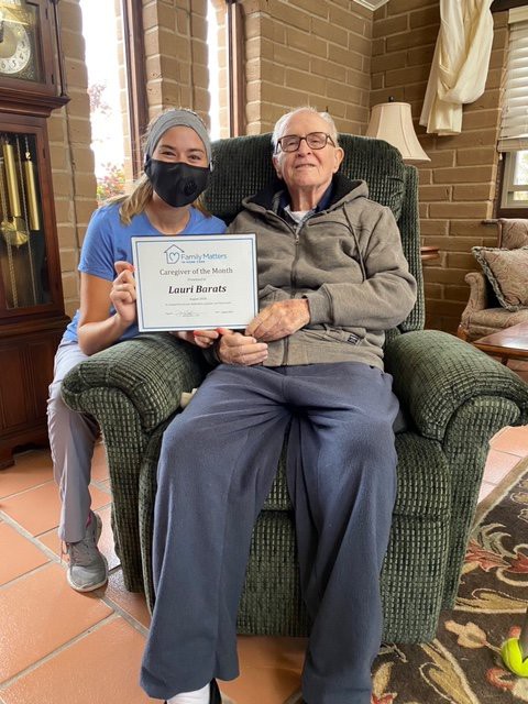 An elderly man seated in a comfortable chair next to a caregiver holding a certificate.