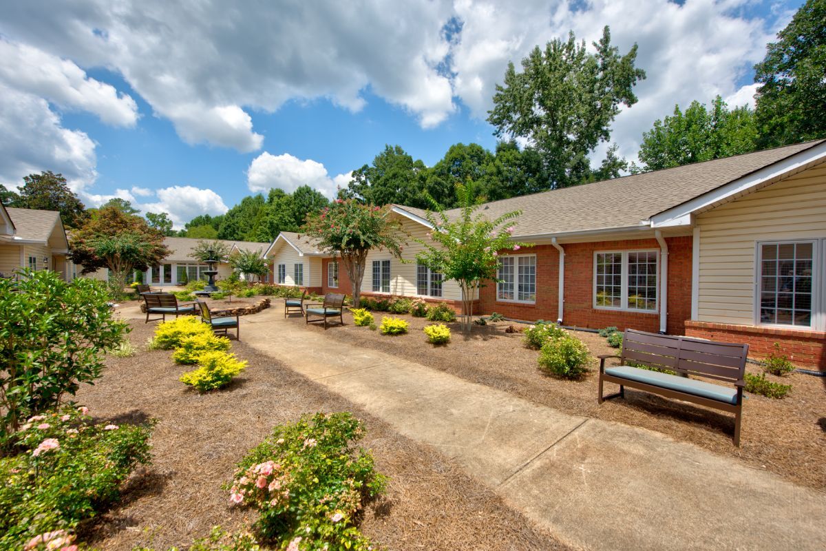A well-maintained garden pathway lined with benches and greenery in front of a brick building under a partly cloudy sky.