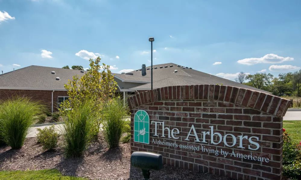 A brick sign for 'The Arbors' in front of a building on a sunny day.