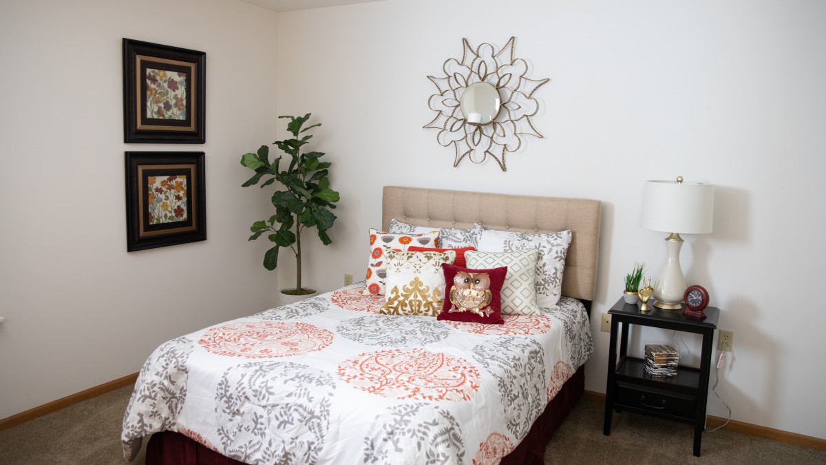 A stylish bedroom with a neatly made bed, decorative pillows, wall art, a sunburst mirror, and a potted plant.
