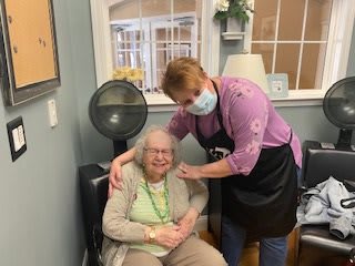 A caregiver embraces an elderly woman seated in a hair salon setting.