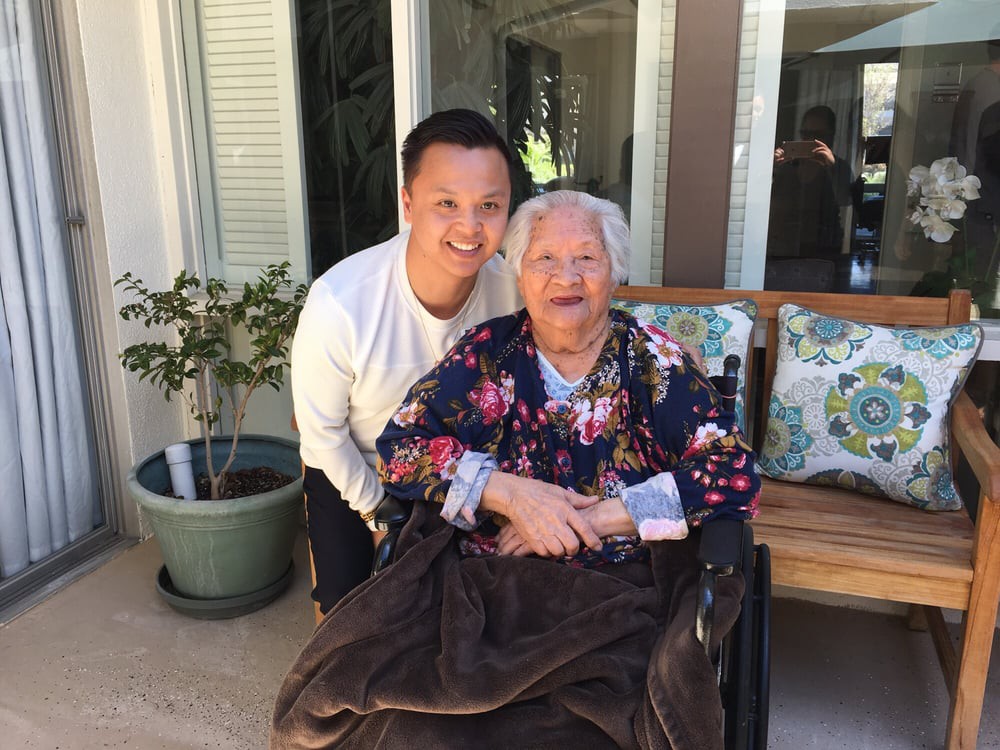 A young man poses happily with an elderly woman seated in a wheelchair, surrounded by a garden setting.