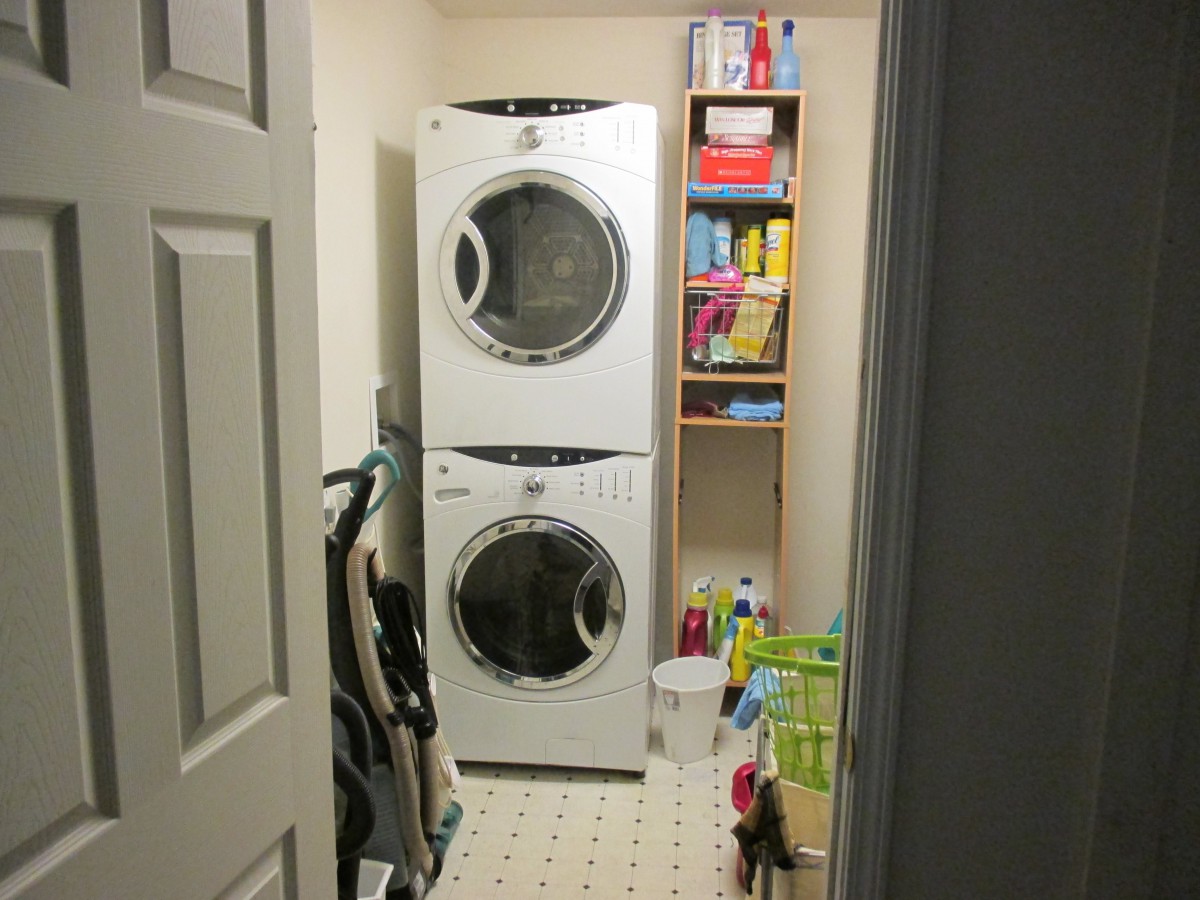 A small laundry room with stacked washer and dryer, storage shelves, and cleaning supplies.