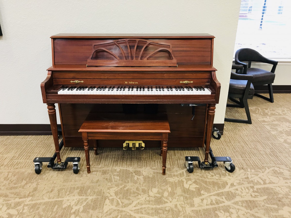 A wooden upright piano with a matching bench, placed on wheels in a music room.