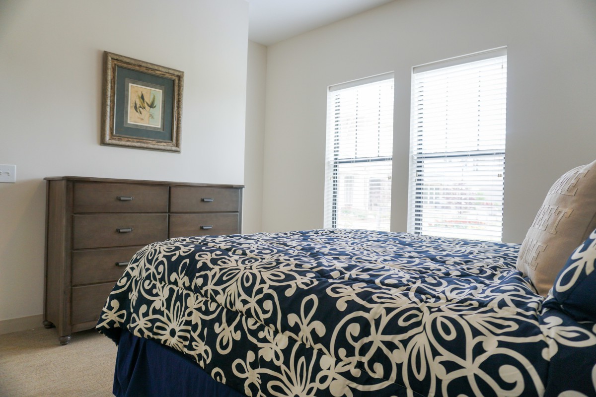 A cozy bedroom featuring a bed with patterned bedding, a wooden dresser, and large windows.
