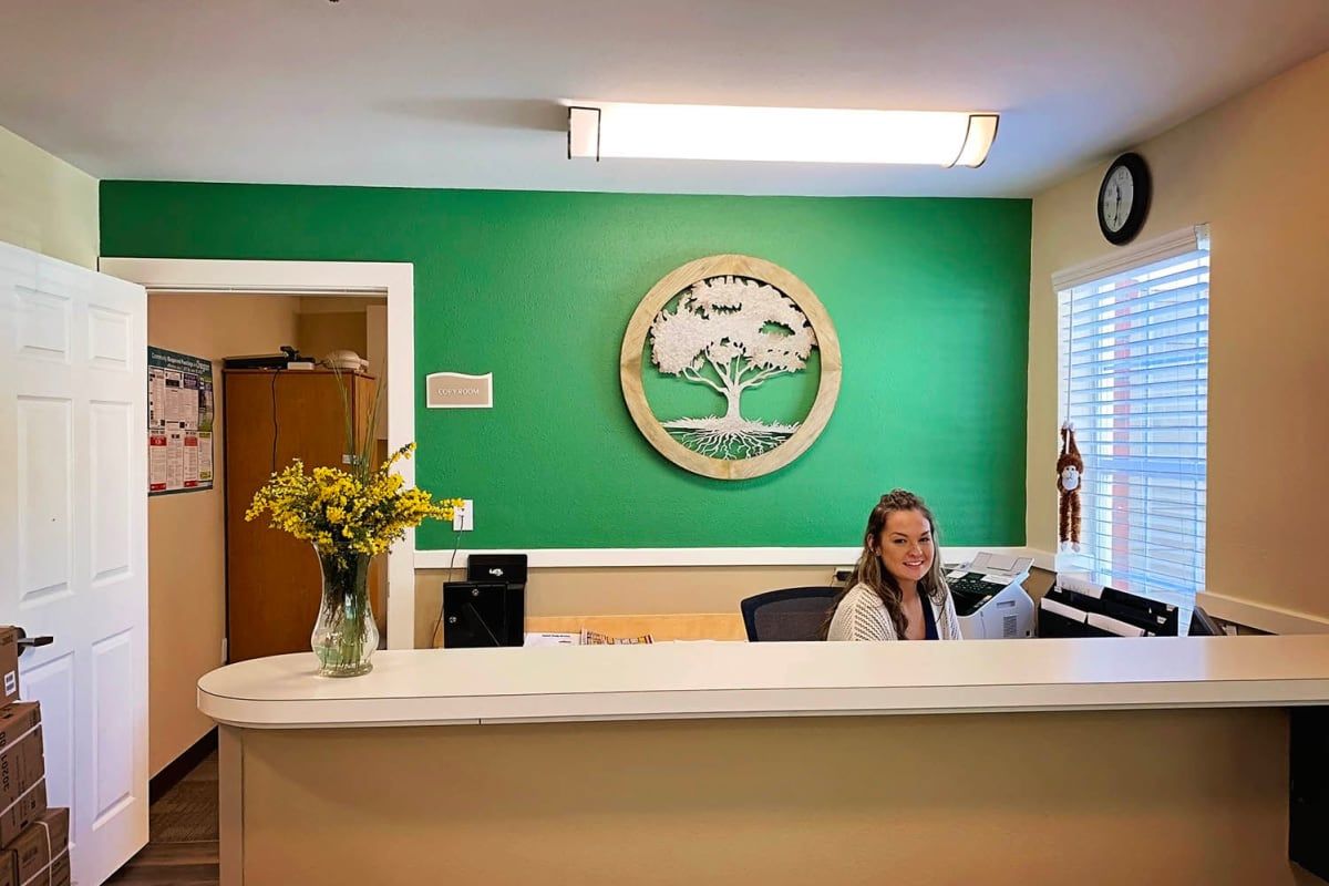 A welcoming reception desk with a woman seated, featuring a green wall and decorative logo.