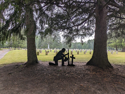 A silhouette of a person kneeling at a grave, surrounded by trees in a serene cemetery.