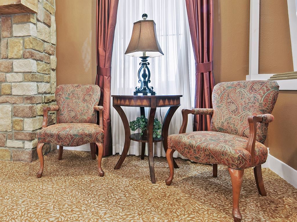 A cozy living room setup with two armchairs flanking a wooden side table and lamp, set against a backdrop of curtains and stone wall accents.