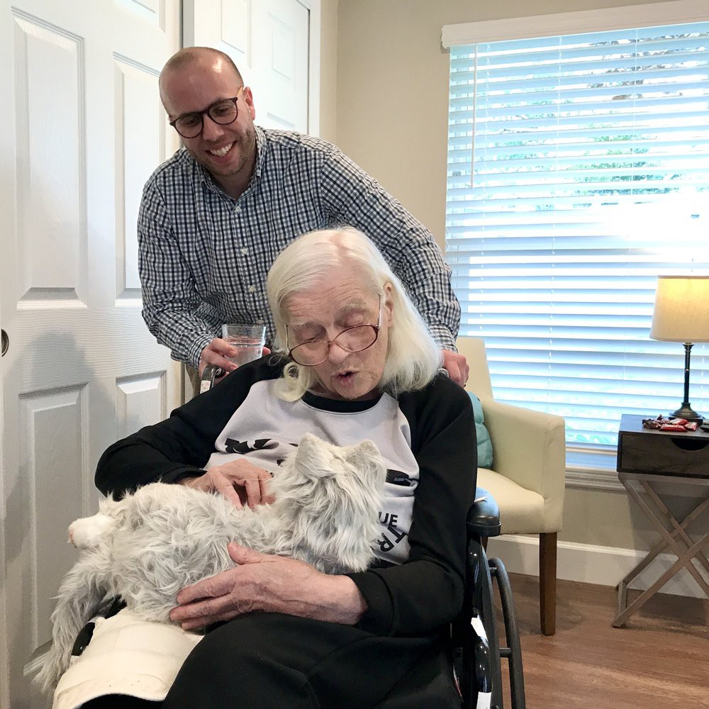 An elderly woman in a wheelchair is holding a dog while a man stands behind her, smiling inside a cozy room.