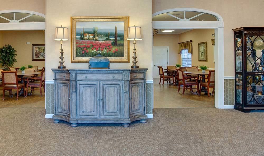 A well-decorated lobby area featuring a wooden cabinet with lamps, a landscape painting, and a dining area in the background.