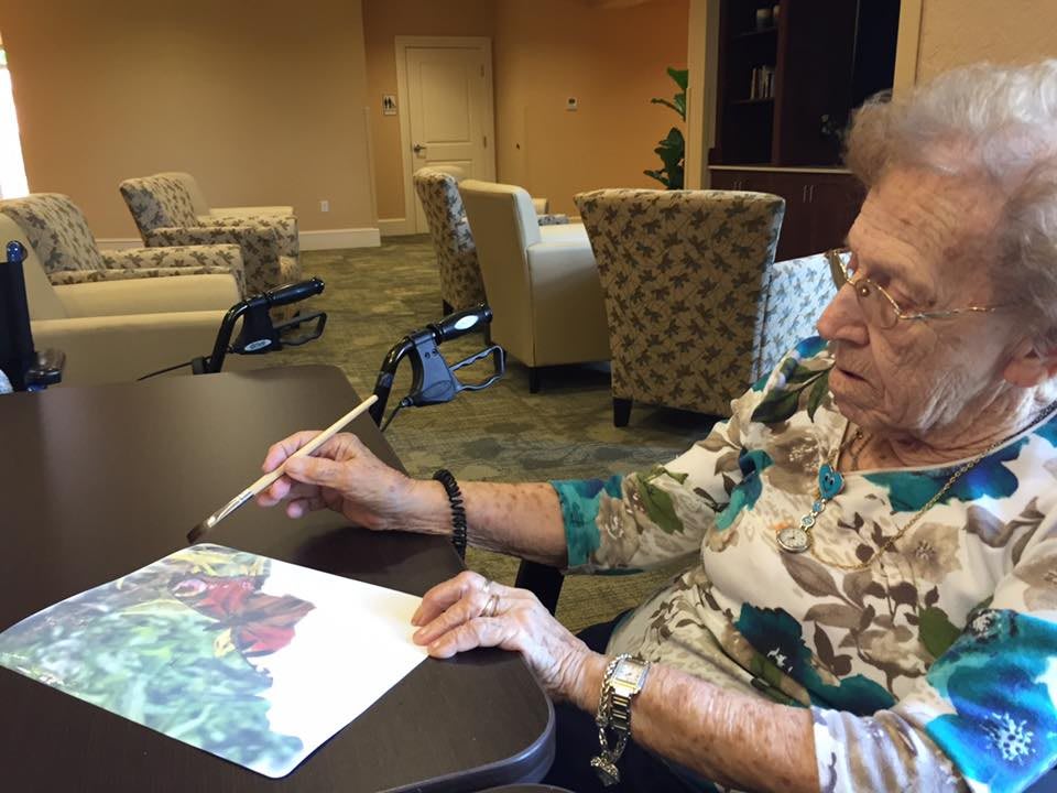An elderly woman is engaged in painting at a table indoors, showcasing her creativity and passion for art.