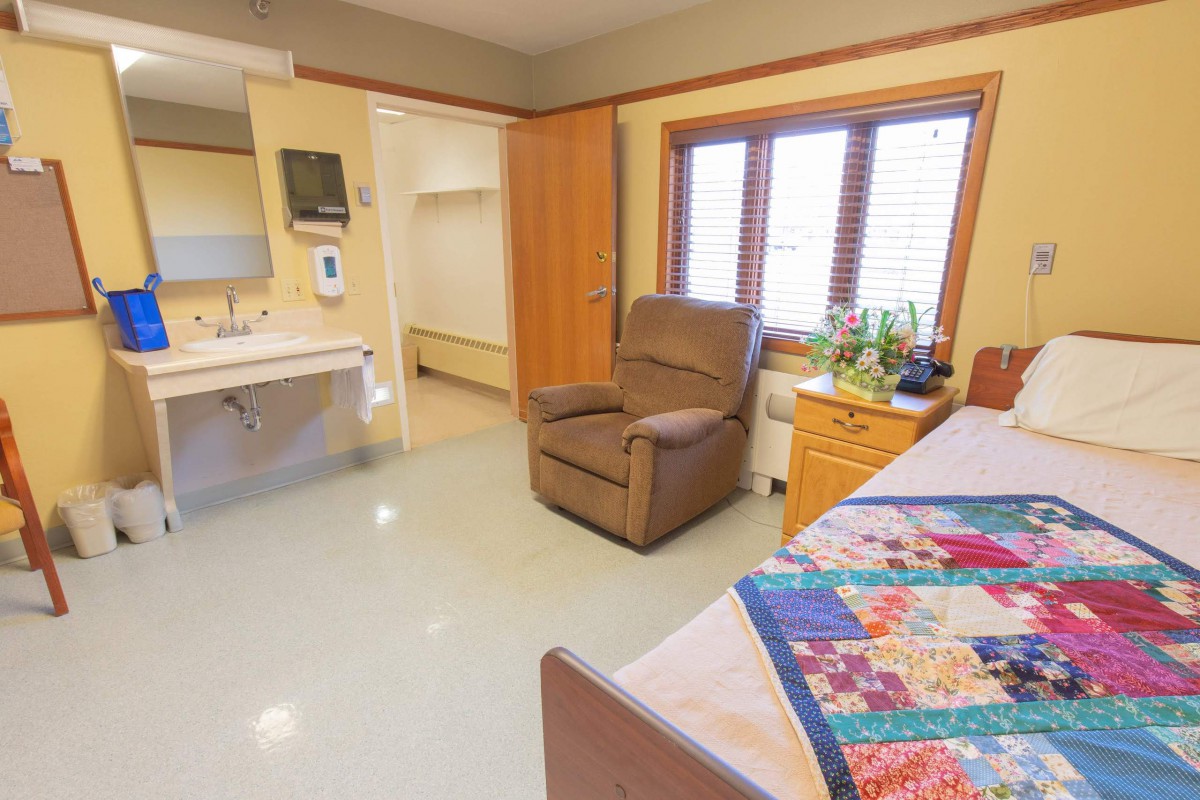 A cozy room with a recliner chair, bed with colorful quilt, and large window.