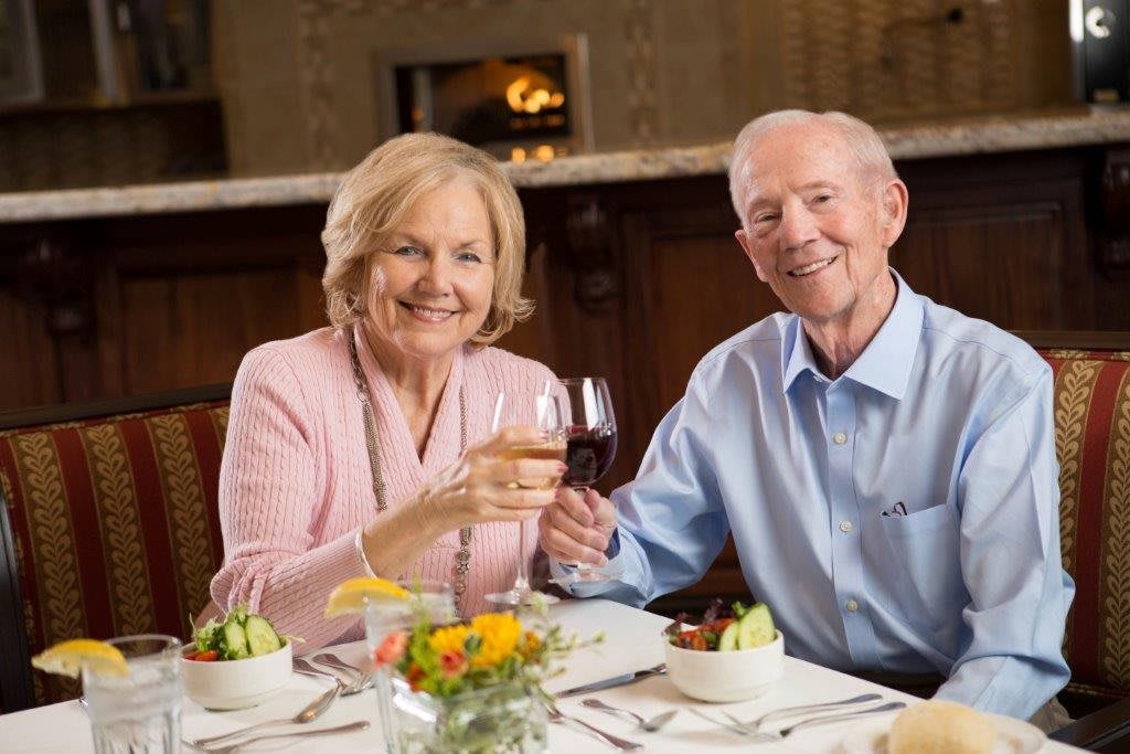 An elderly couple smiling and toasting with wine at a restaurant table.