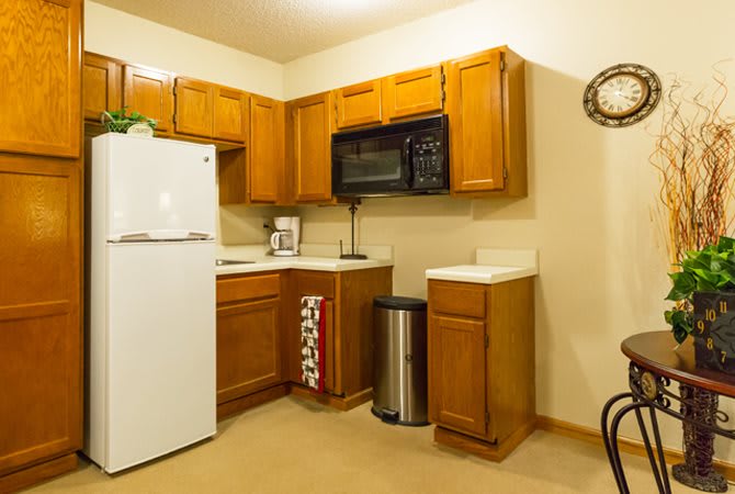 A cozy kitchen featuring wooden cabinets, a refrigerator, microwave, and a trash can.