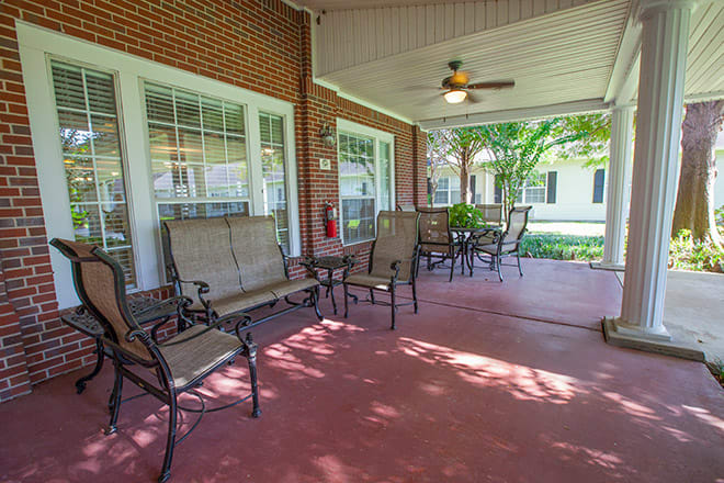 A cozy outdoor patio area with seating, tables, and a ceiling fan, surrounded by brick walls and windows.