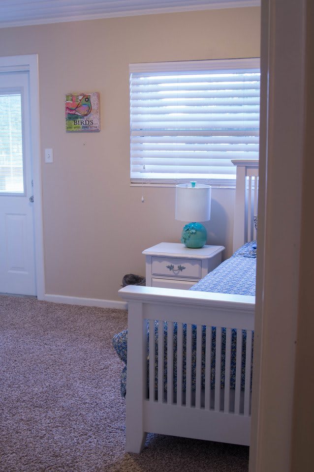 A cozy bedroom with a white nightstand, lamp, and a bed next to a window.