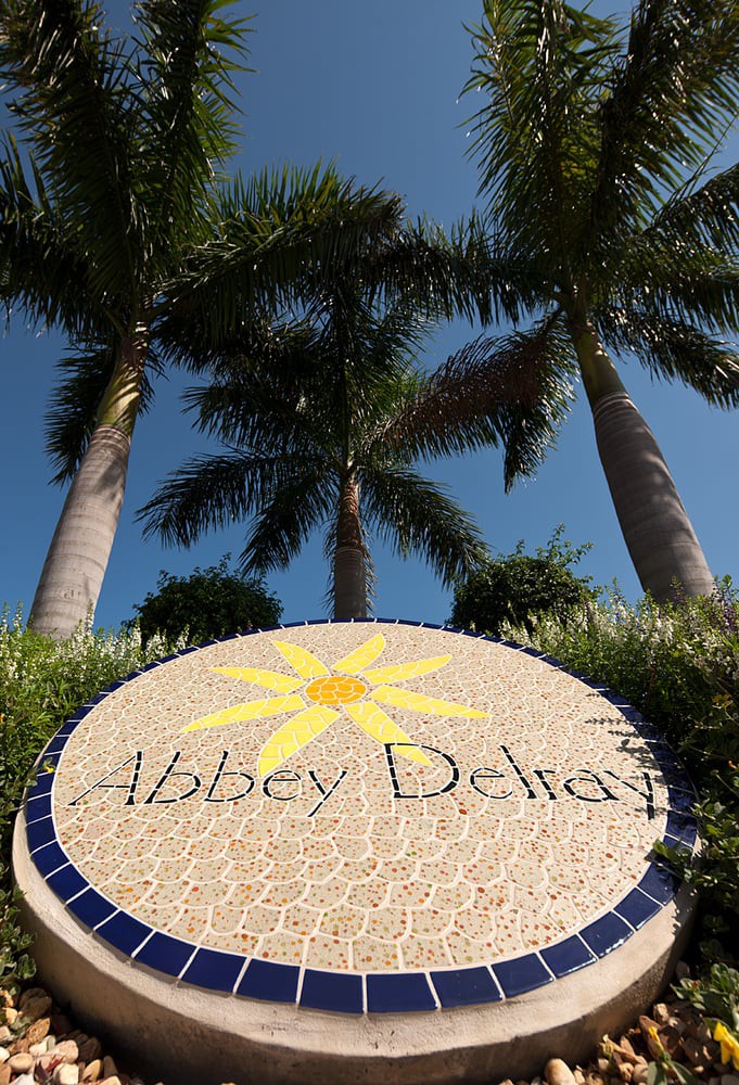 A mosaic sign with 'Abbey Delray' depicted, set against towering palm trees and a clear blue sky.