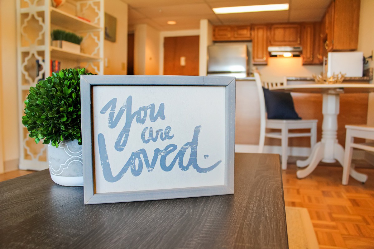 A framed sign in a cozy home setting reads 'You are loved' in front of a kitchen area.