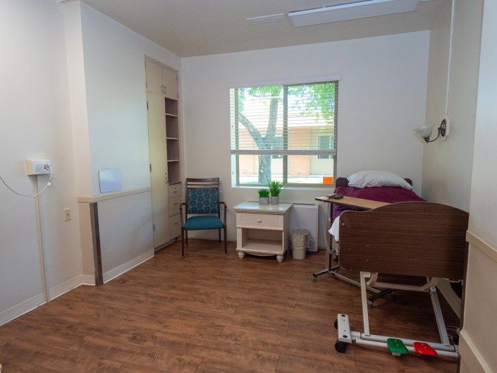 A well-lit room featuring a hospital bed, chair, and storage shelves with a view of the outside through a window.