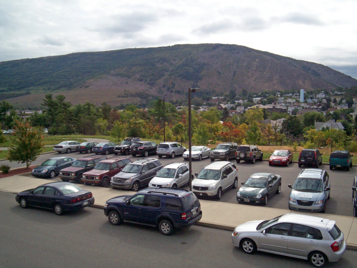 A parking lot filled with cars against a backdrop of hills and a cloudy sky.