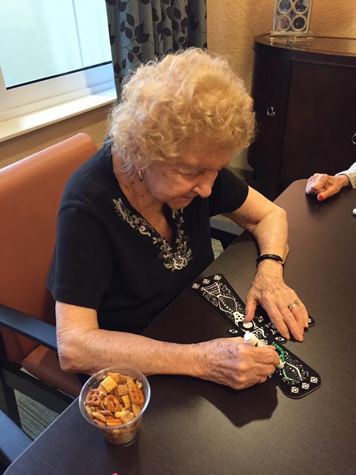 An elderly woman engaged in a game of dominoes at a table with snacks nearby.
