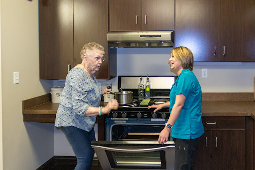 An elderly woman and a caregiver engage in conversation while preparing a meal in the kitchen.