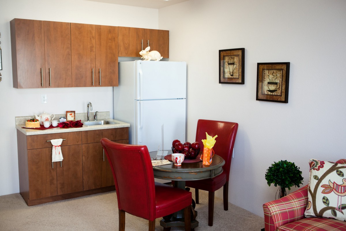 A cozy kitchen and dining area featuring a small table with red chairs, wooden cabinets, and a fridge.