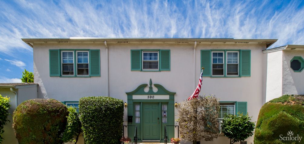 A charming white house with green shutters and a door, displaying an American flag, under a partly cloudy sky.