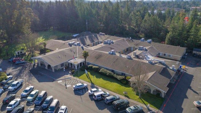 Aerial view of a large building complex surrounded by trees, with a parking lot filled with cars.