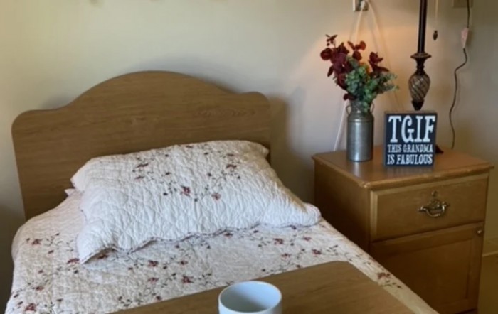 A cozy bedroom scene with a floral patterned bedspread, a vase of flowers on the nightstand, and a small decorative sign.