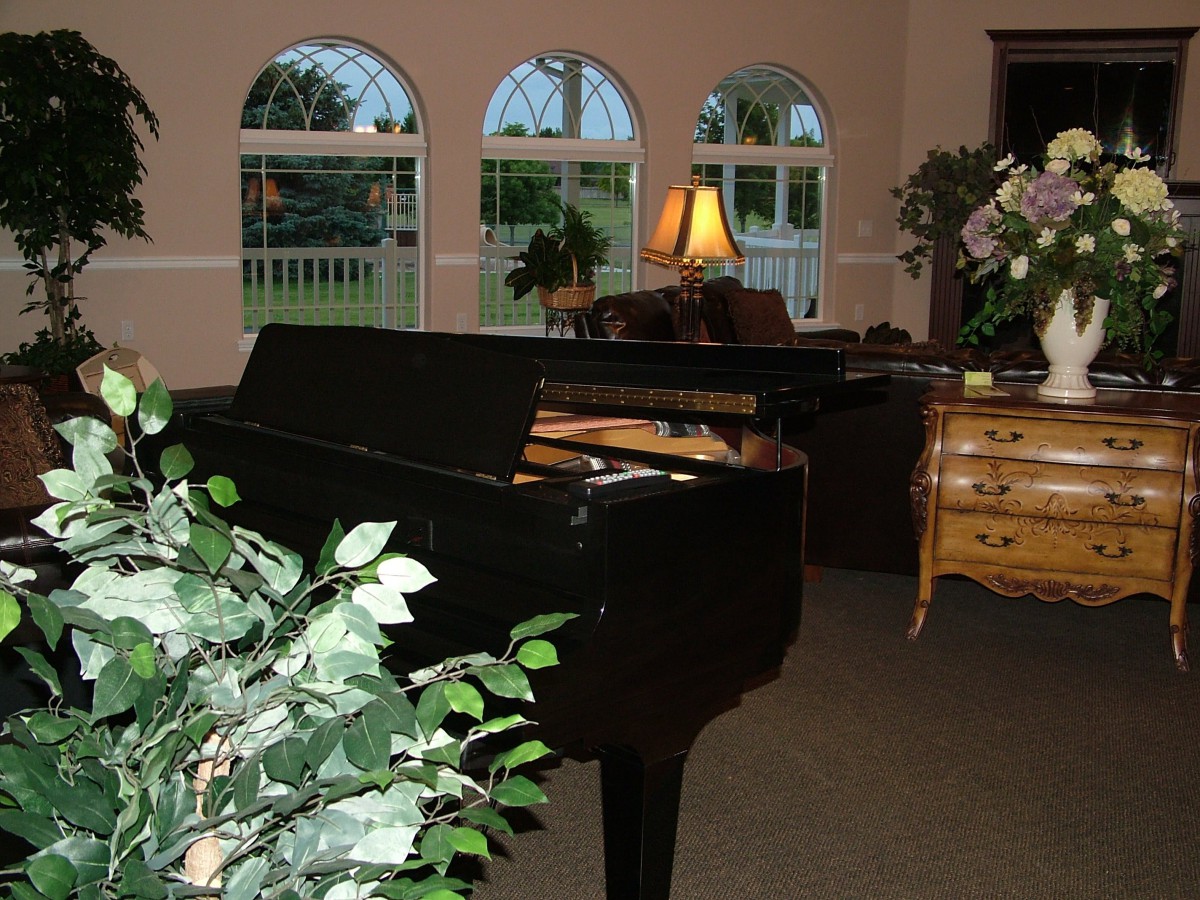 A cozy living room featuring a grand piano, with plants and warm lighting from a lamp next to a set of arched windows.