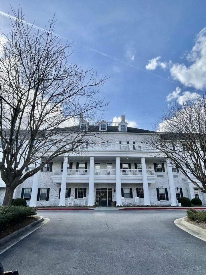 A colonial-style building with a white facade and large columns, set against a backdrop of trees and a cloudy sky.