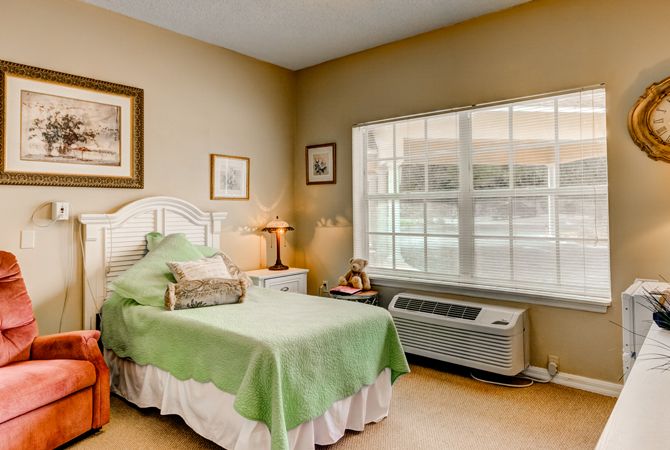 A cozy bedroom with a green bedspread, framed artwork, and large windows.