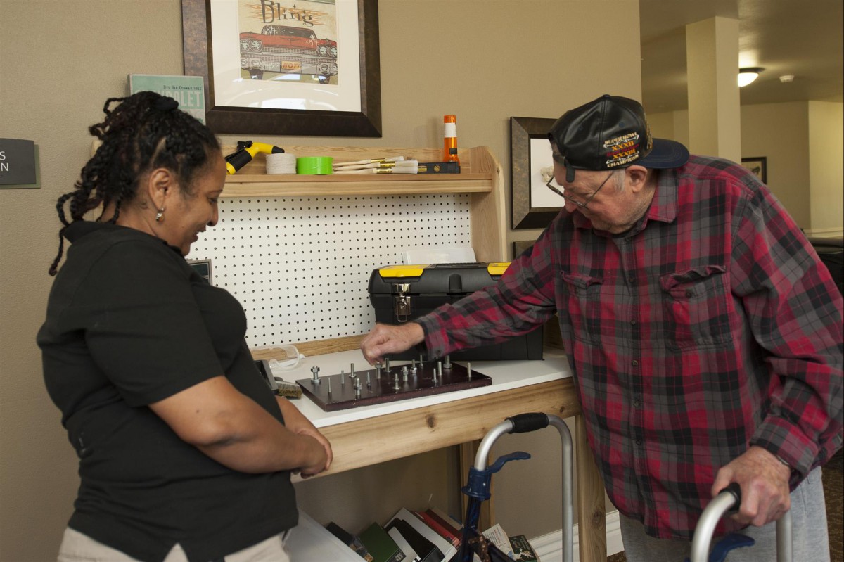 An elderly man and a woman interact by a workbench with tools in an indoor setting.