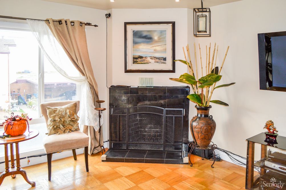 A cozy living room corner featuring a framed artwork above the fireplace, a potted plant, and a comfortable chair by the window.