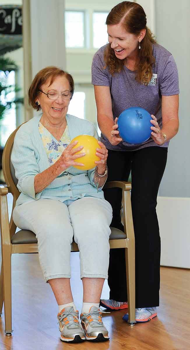 A senior woman participates in a physical therapy session with the assistance of a trainer, using medicine balls for exercise.