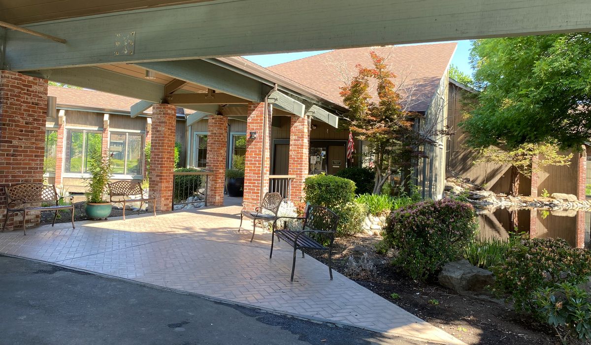 A sunny day view of a building entrance with chairs and landscaping.